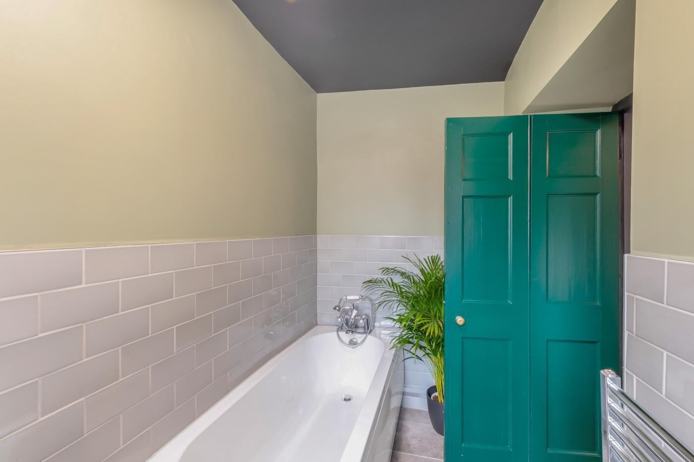 A bathroom with a bathtub, grey and white tiled walls, a green door, and a potted plant.