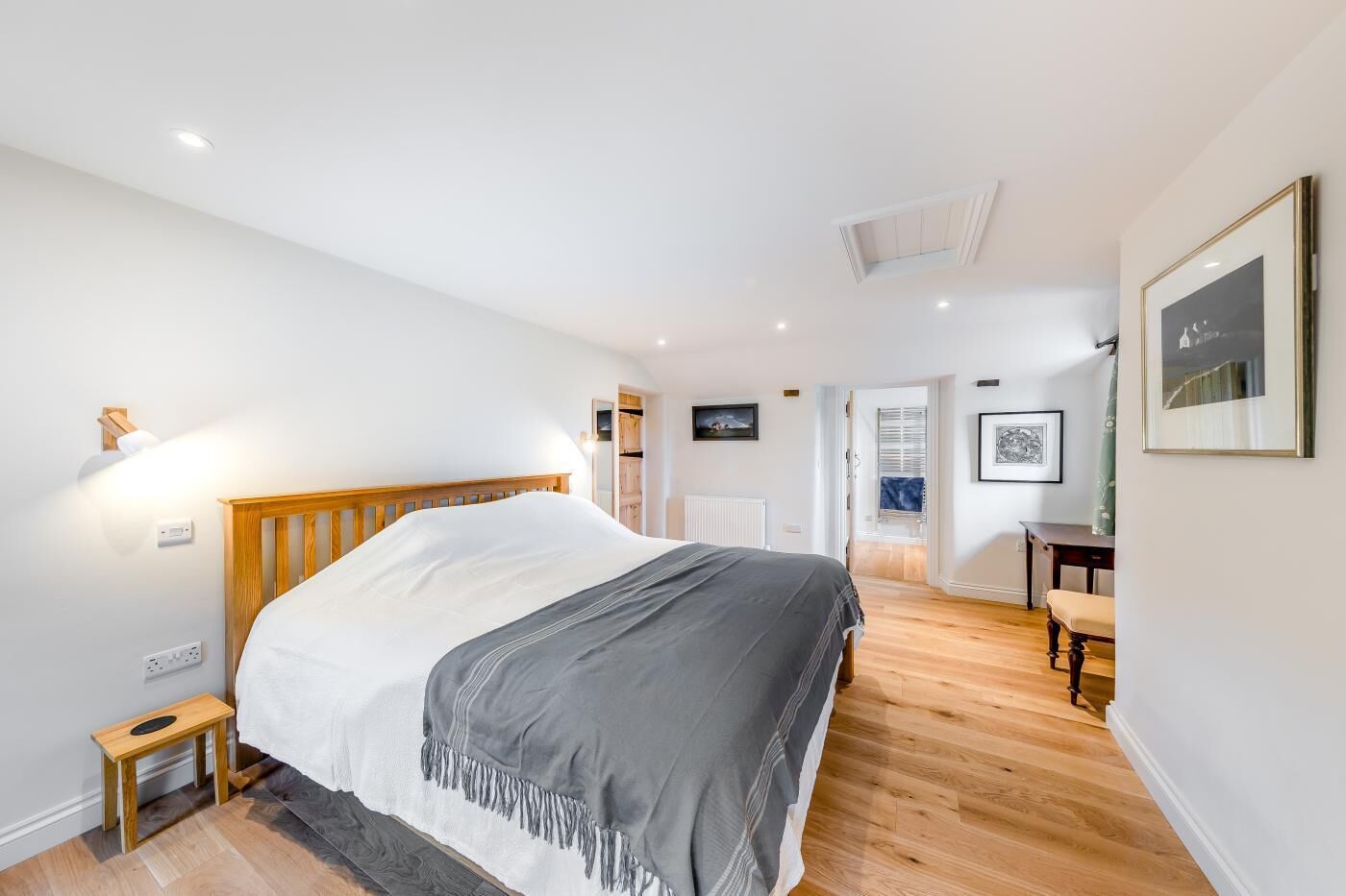 A bedroom with a wooden bed, bedside table, and a small stool. There is a door leading to another room and a window with blinds.