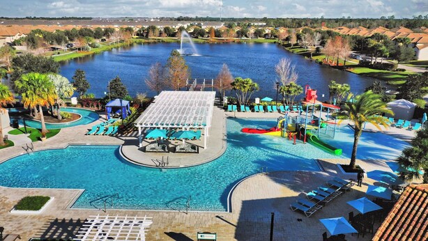 Une grande piscine extérieure avec un toboggan, entourée de chaises longues et de parasols, et offrant une vue sur un lac et un quartier résidentiel en arrière-plan.