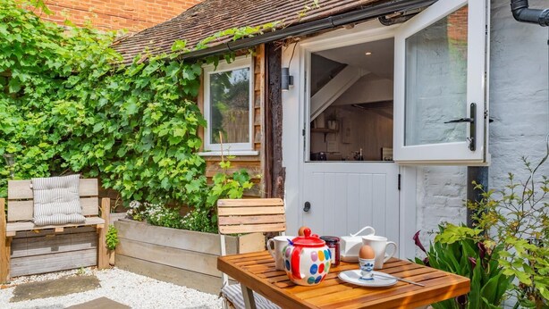 A cozy outdoor seating area with a wooden table and bench, a white door, and a brick wall with green ivy. - 1 Bed in Chilham (oc-t29281) (Chilham)
