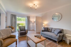 A spacious living room with a sofa, armchair, and coffee table, a large mirror, and a view of greenery outside. - 3 Bed in Tenby (oc-o29381) (Tenby)