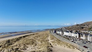 A coastal town with a beach, a road lined with buildings, and a clear blue sky. - 3 Bed in Dyffryn Ardudwy (oc-f29350) (Dyffryn Ardudwy)
