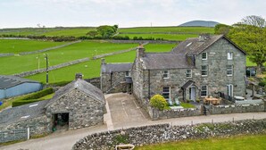 A stone building with a paved courtyard, a small attached structure, and a grassy field with sheep in the background. - 3 Bed in Dyffryn Ardudwy (oc-f29350) (Dyffryn Ardudwy)