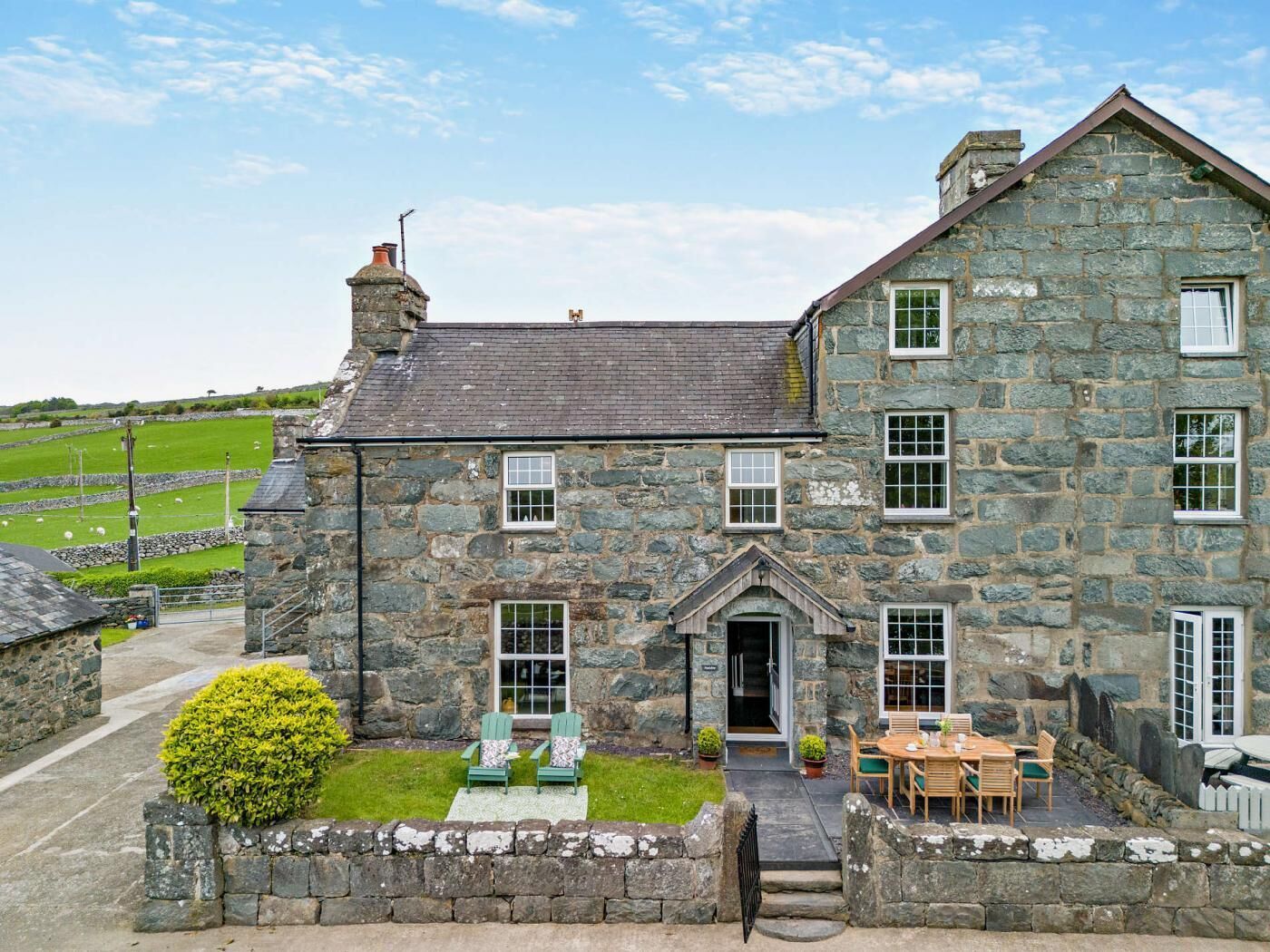 A stone house with a traditional design, featuring a front porch with two chairs and a table, a stone wall surrounding the property, and a clear view of the surrounding landscape.