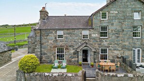 A stone house with a traditional design, featuring a front porch with two chairs and a table, a stone wall surrounding the property, and a clear view of the surrounding landscape. - 3 Bed in Dyffryn Ardudwy (oc-f29350) (Dyffryn Ardudwy)