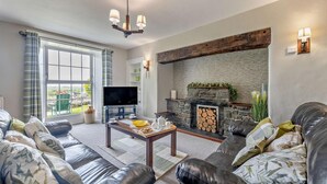 A modern living room with a stone fireplace, leather sofas, a flat-screen TV, and a large window offering a view of the outdoors. - 3 Bed in Dyffryn Ardudwy (oc-f29350) (Dyffryn Ardudwy)