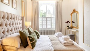 Ein Schlafzimmer mit einer gepolsterten Kopfteile, einem Nachttisch mit Lampe, einem großen Spiegel und Blick auf ein Gebäude durch ein Fenster.