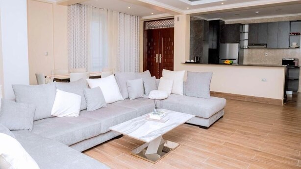 A modern living room with a grey sectional sofa, a white coffee table, and a kitchen area with dark cabinets and a microwave. - One-Bedroom Apartment #9 (Kigali)
