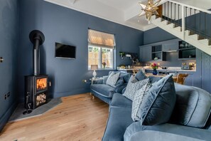 Un salon moderne doté d’un poêle à bois encastré, d’un canapé gris agrémenté de coussins à motifs, d’un plancher en bois et d’un escalier menant à un niveau supérieur.