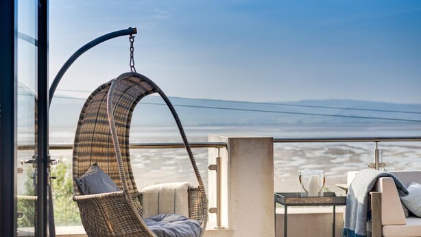 A balcony with a hanging chair, a small table with a pitcher, and a view of the ocean. - 5 Bed in Aberdovey (oc-t29402) (Aberdovey)