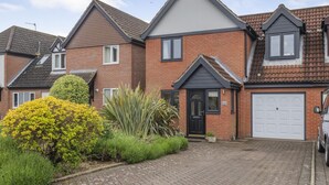 Een woonhuis van baksteen met twee verdiepingen, een aangebouwde garage, een zwarte voordeur en een kleine tuin met gele en groene planten.