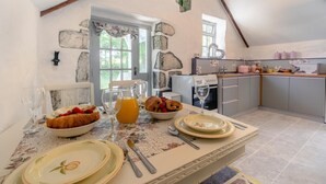 A breakfast setup with croissants, fruit, and orange juice on a table with a floral tablecloth. A kitchen with a stone wall and a window is visible in the background. - 2 Bed in Trewoon (oc-t29504) (Saint Mewan)