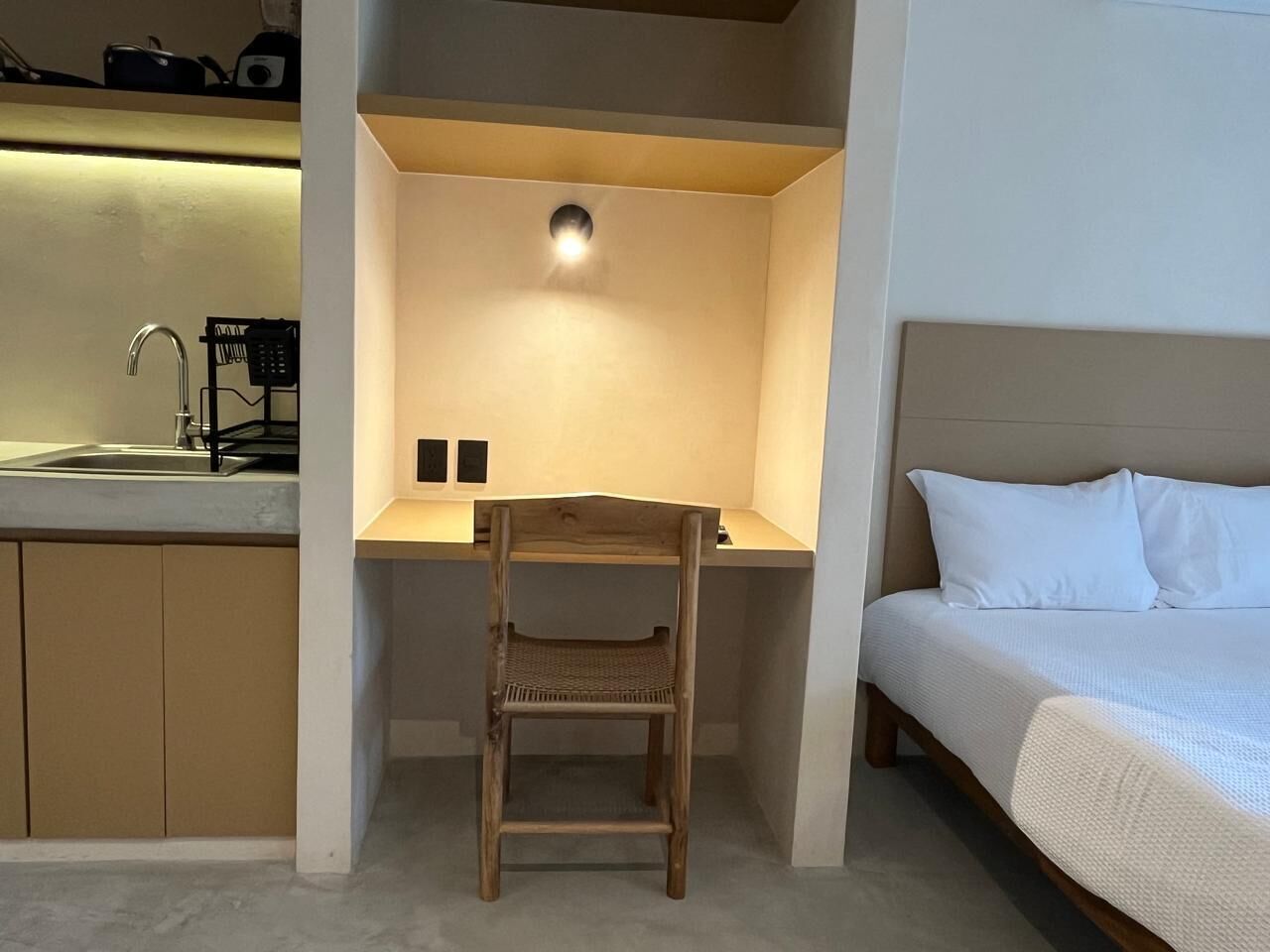 A compact room with a kitchenette, a wooden chair, a bed, and a small desk area with a light fixture.