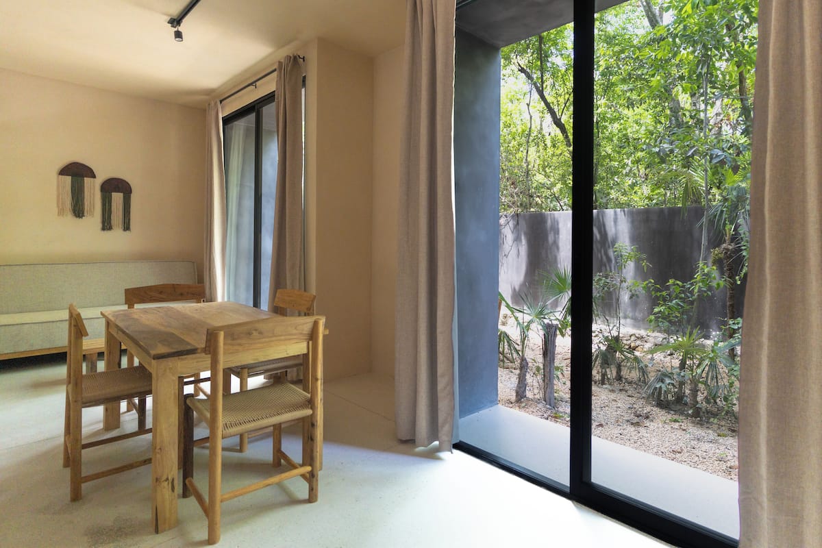 Quarto com mesa e cadeiras de madeira, uma janela grande com cortinas e vista para um jardim.