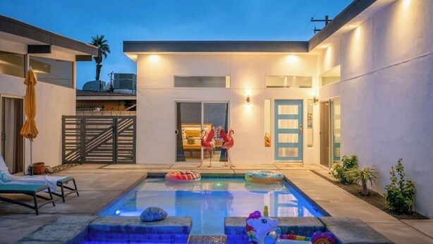 Un área de piscina moderna al aire libre con sillas de descanso, una piscina con juguetes flotantes y un edificio de dos pisos con una puerta azul.