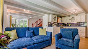 A living room with a blue sofa and armchair, a wooden staircase, and a kitchen with white cabinets and a blue countertop.