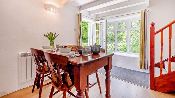 A dining area with a wooden table and chairs, a vase of flowers, and a view of the outdoors through large windows.