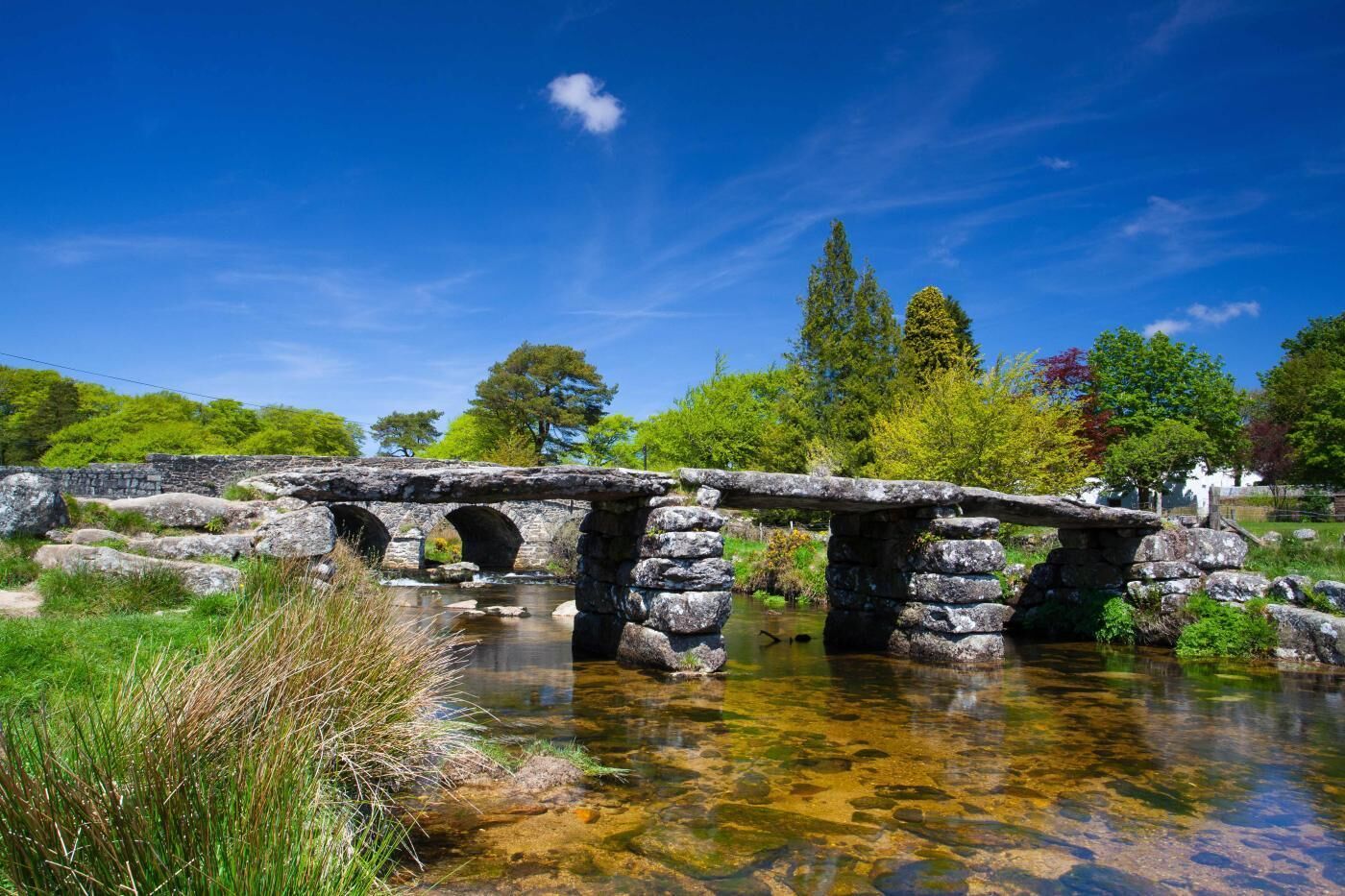 A stone bridge over a clear stream with surrounding greenery and trees.