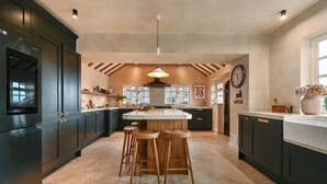 Een moderne keuken met donkere kastjes, een centraal eiland met krukken, hanglampen en een grote klok aan de muur.