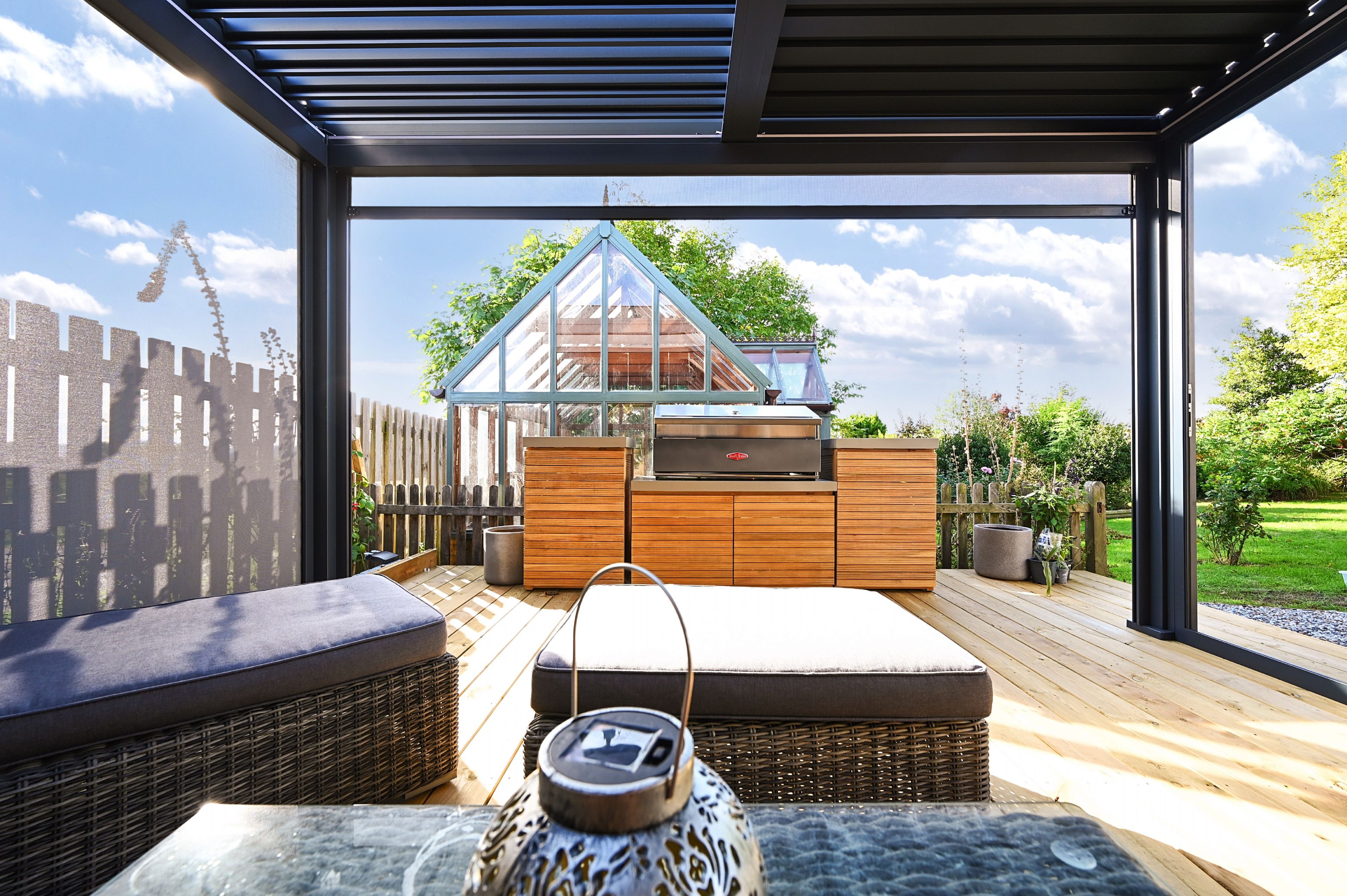 Un área de estar moderna al aire libre con cerramiento de vidrio, terraza de madera y vista a una casa con techo verde.