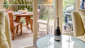 Una mesa de vidrio con una botella de vino y dos copas, una mesa de madera con sillas, y un balcón con vistas a árboles.