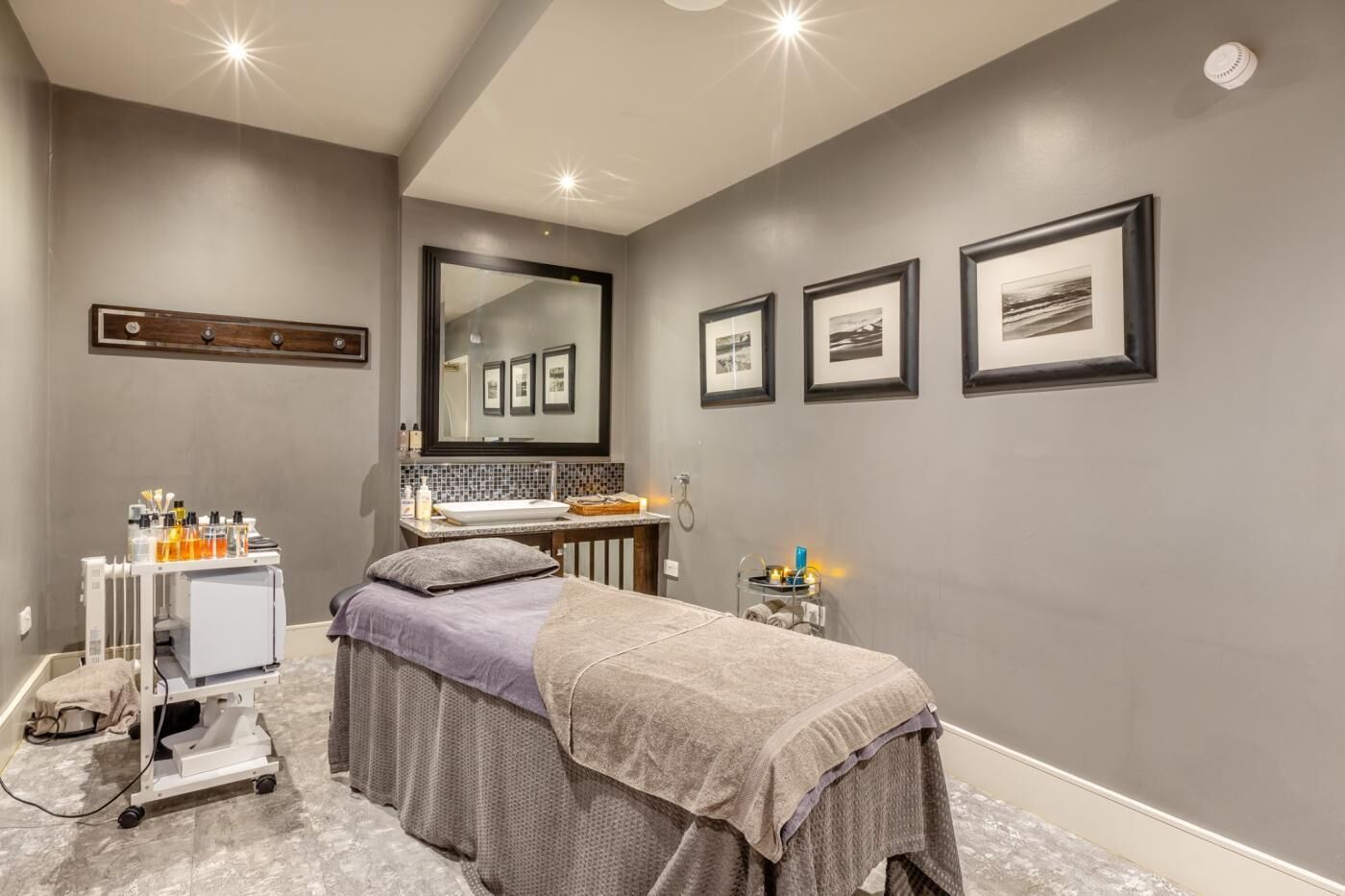 A modern spa room with a massage table, a vanity with a mirror, and a small shelf with toiletries.