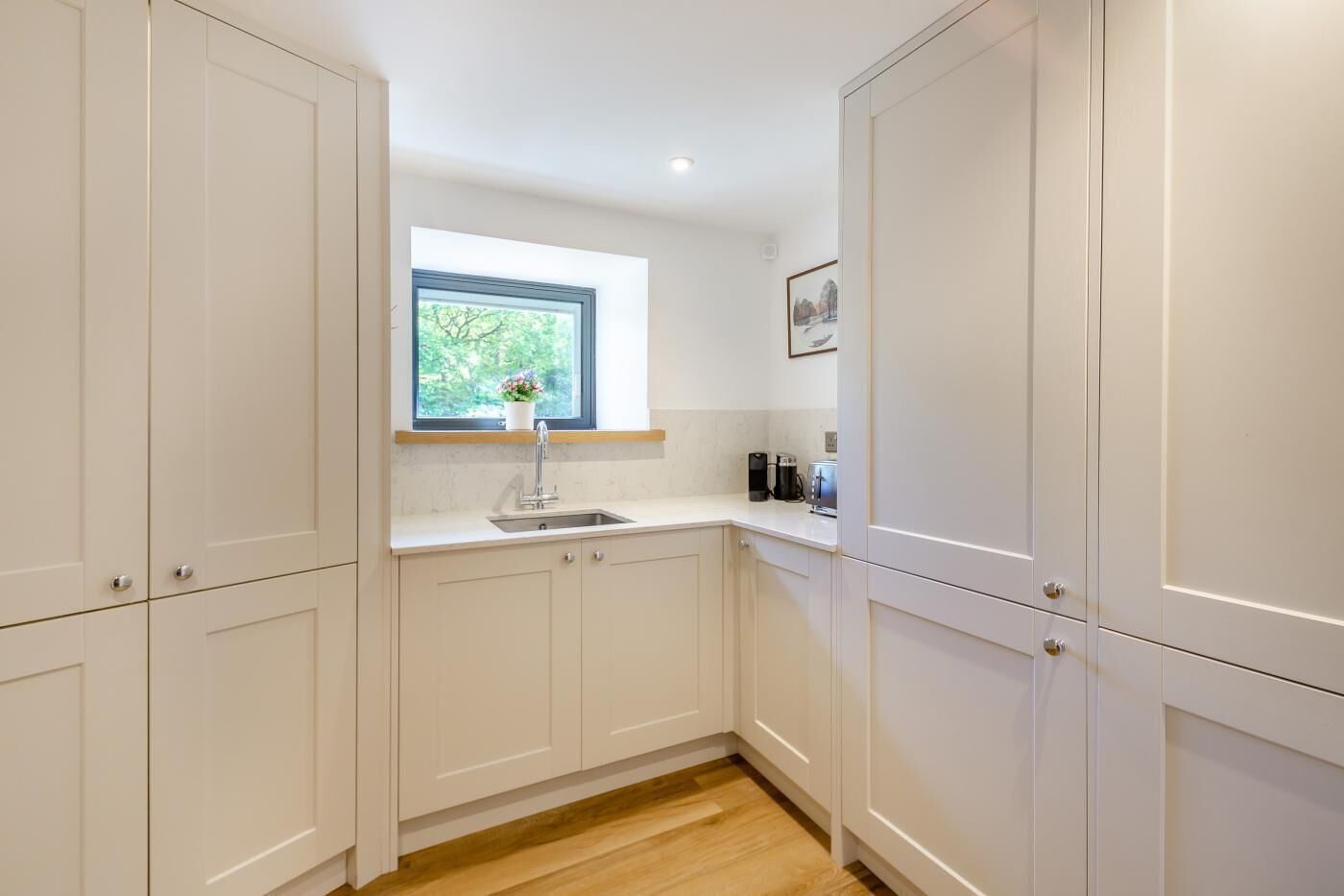 Una cocina moderna con armarios blancos, una ventana con vista a la vegetación, un lavabo y una encimera con cafetera.