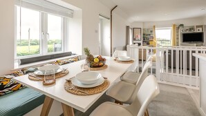 A dining area with a white table set for four, a view of the outdoors through large windows, and a living area with a TV and bookshelves. - 2 Bed in St. Minver (oc-r29475) (Saint Minver)
