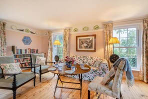 Ein gemütliches Zimmer mit einer Couch, einem Tisch mit Blumenvase, einem Bücherregal und einem Fenster mit Vorhängen.