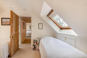 Ein Schlafzimmer mit einem Oberlicht, einer Holztür, einem Bett, einem kleinen Tisch mit einer blumigen Tischdecke und einem gerahmten Bild an der Wand.