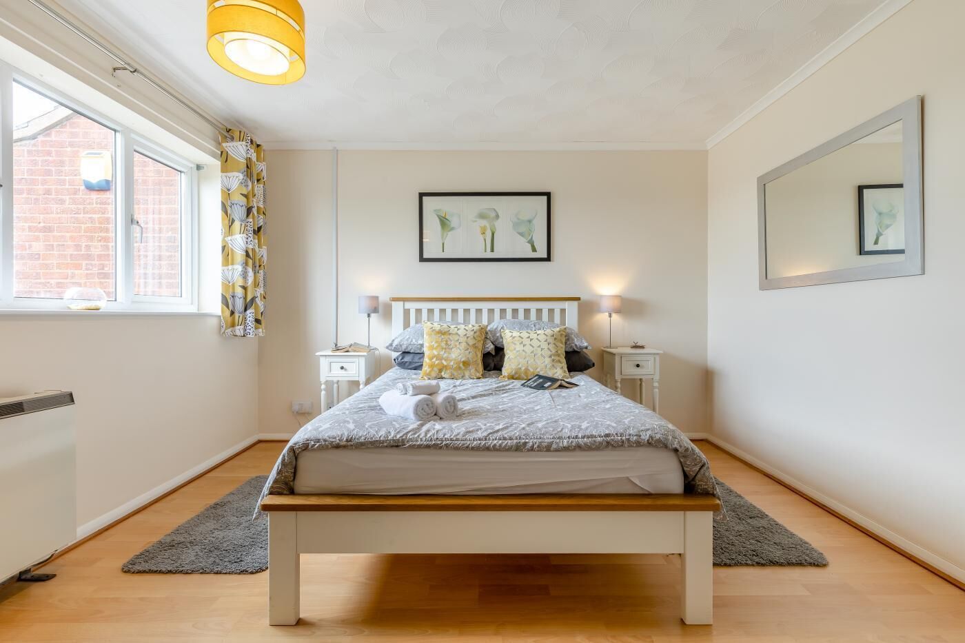 A bedroom with a bed, bedside tables, a radiator, a window with curtains, and artwork on the walls.