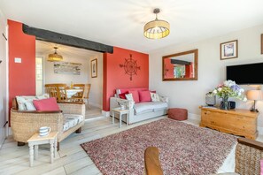 A cozy living room with a red accent wall, a white sofa, wicker chairs, a wooden coffee table, and a large rug. - 2 Bed in Reedham (oc-q29527) (Haddiscoe)
