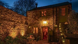 A stone house with a well-lit entrance, a small patio with a table and chairs, and a garden area with plants and flowers. - 1 Bed in Reeth (oc-t29540) (Reeth)
