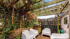 A covered outdoor seating area with white plastic chairs, a brick wall, and a wooden pergola with string lights. - 2 Bed in Rye (oc-86094) (Rye)