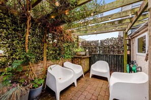 A covered outdoor seating area with white plastic chairs, a brick wall, and a wooden pergola with string lights. - 2 Bed in Rye (oc-86094) (Rye)