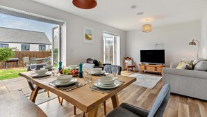 A spacious living room with a wooden dining table set for four, a grey sofa, a television, and a fireplace. - 3 Bed in Newquay (oc-blueb6) (Newquay)