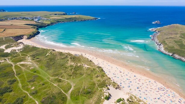 An aerial view of a coastal area with a beach, clear blue waters, and green fields. - 3 Bed in Newquay (oc-blueb6) (Newquay)