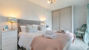 A modern bedroom with a large bed, two bedside lamps, a nightstand with a picture frame, and a bench with a cushion. - 3 Bed in Newquay (oc-blueb6) (Newquay)