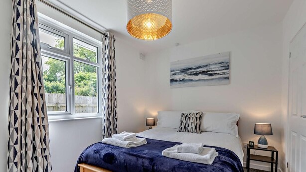 Ein Schlafzimmer mit einem großen Bett, zwei Nachttischlampen, einem Fenster mit gemusterten Vorhängen, einem Wandbild und einer deckenhohen Leuchte mit Struktur.