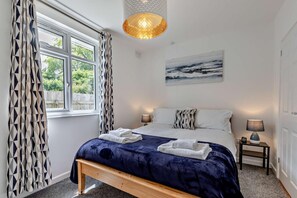 Quarto com cama grande, duas luminárias de cabeceira, janela com cortinas estampadas, quadro na parede e luminária de teto com textura.