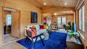 Interior acolhedor de uma cabana com decoração em madeira, tapete azul, sofá branco com almofadas coloridas, área de refeições e uma janela grande.