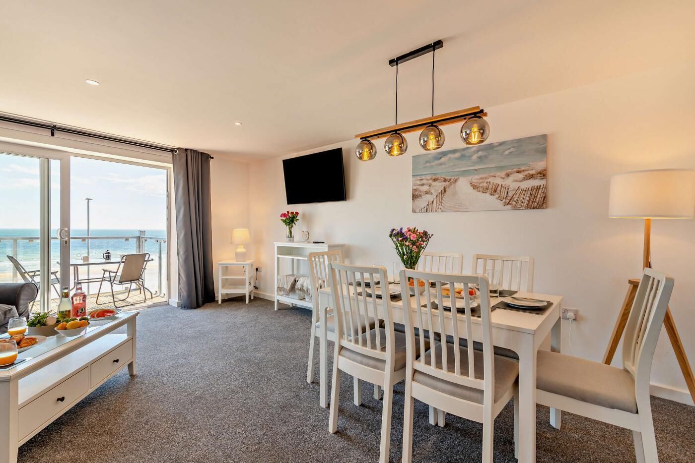 A hotel room with a balcony, a dining area with white furniture, a flat-screen TV mounted on the wall, and a painting of a beach scene.