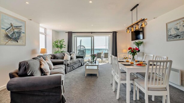 Sala de estar moderna com sofá, mesa de jantar e cadeiras. Há vista para o mar através de grandes janelas.