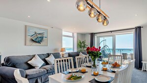 Uma sala de estar moderna com um sofá branco, uma mesa de jantar posta com pratos e copos, e vista para o mar através de grandes janelas.