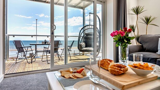 A room with a balcony overlooking the sea, featuring a dining table with food, a sofa, and a vase of flowers.