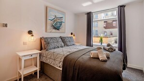 A bedroom with a bed, a nightstand, a painting, a window with curtains, and a view of buildings outside.