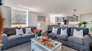A modern living room with two grey sofas, a coffee table with food, and a view of the kitchen and outside.