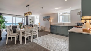 A modern kitchen with grey cabinets, a dining area with white chairs, and a view of the sea through large windows.