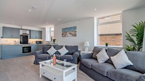 Ein modernes Wohnzimmer mit grauen Sofas, einem Glastisch und einer Küche im Hintergrund.