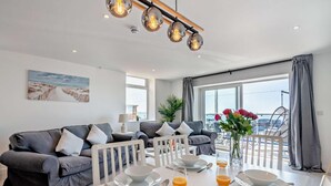 Ein modernes Wohnzimmer mit einer grauen Sofagarnitur, einem Esstisch für zwei Personen und einem großen Fenster mit Meerblick.